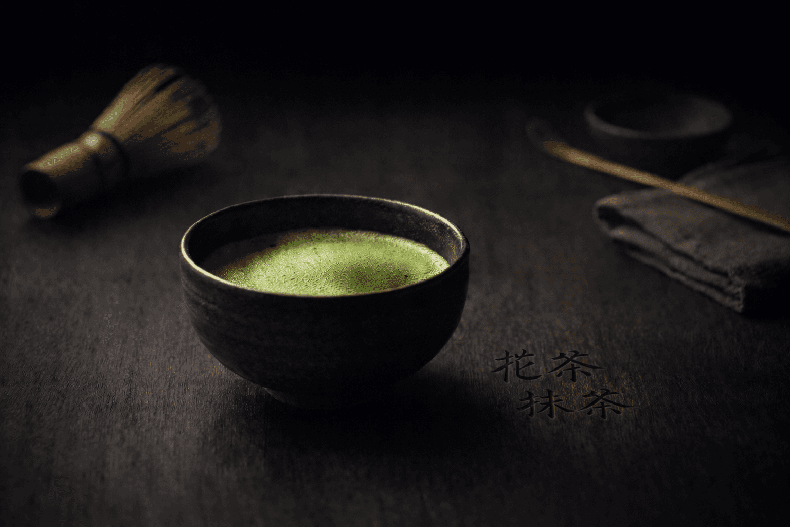 A bowl of matcha resting on a dark timber surface with preparation tools nearby